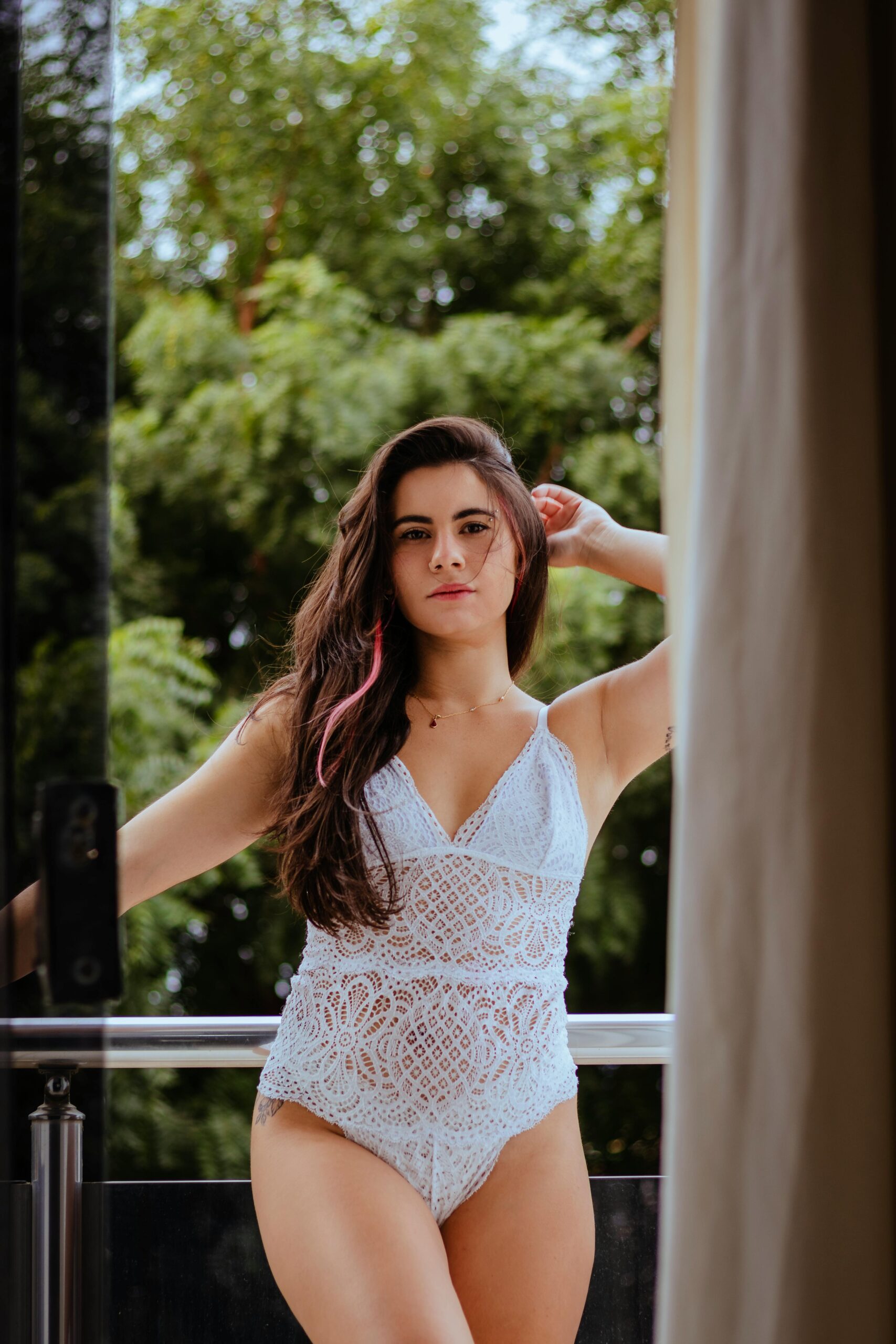 Woman in a lace outfit posing confidently on a balcony with lush greenery in the background.